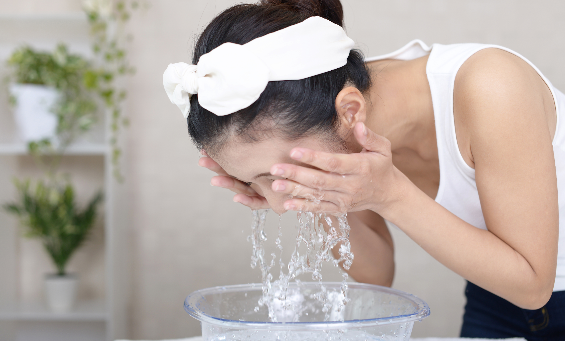 Woman washing her face in a glass sink, with her hair held back by a headband