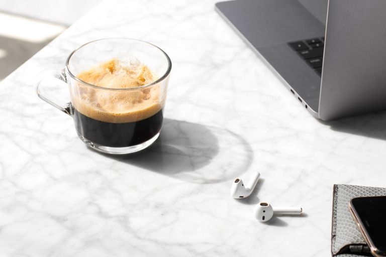 Cup of coffee sitting on a white marble surface with airpods and a laptop nearby