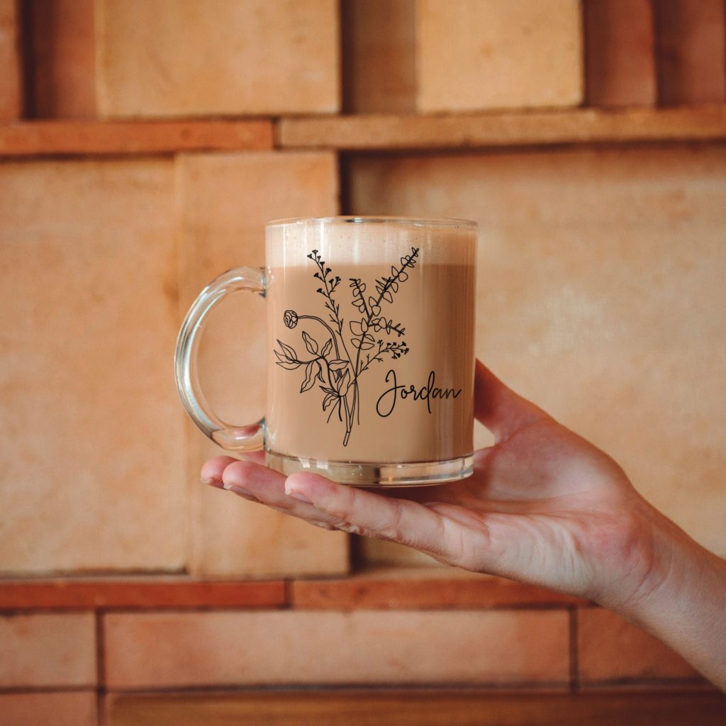 Someone's hand holding a clear glass coffee mug with a floral design and a woman's name etched into it