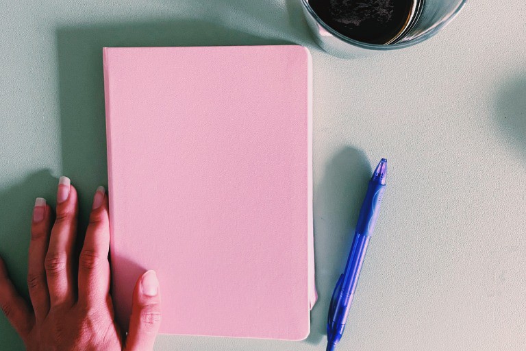 Pink journal sitting on a desk, with a purple pen and a cup of cold brew coffee