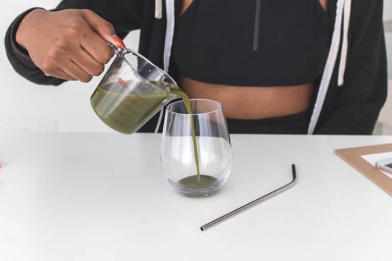 Woman pouring green juice into a glass, with a reusable metal straw lying on the counter