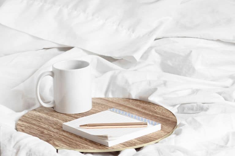 White coffee mug, a pad of paper, and a pen sitting on a wooden round tray on a white bedsheet