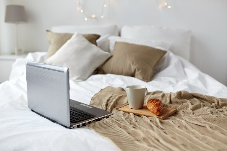 Bed with beige pillows, laptop, a cup of coffee, and a croissant